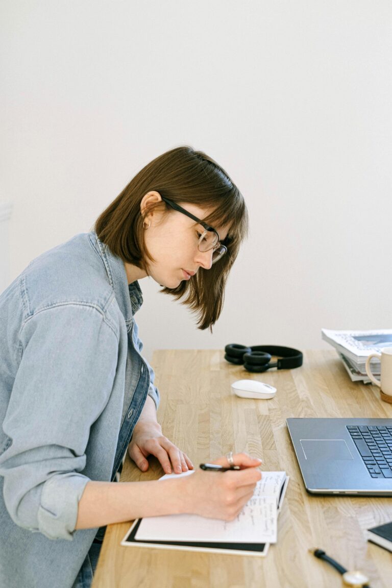 Focused young woman taking notes in a cozy home office setting with laptop and stationery.